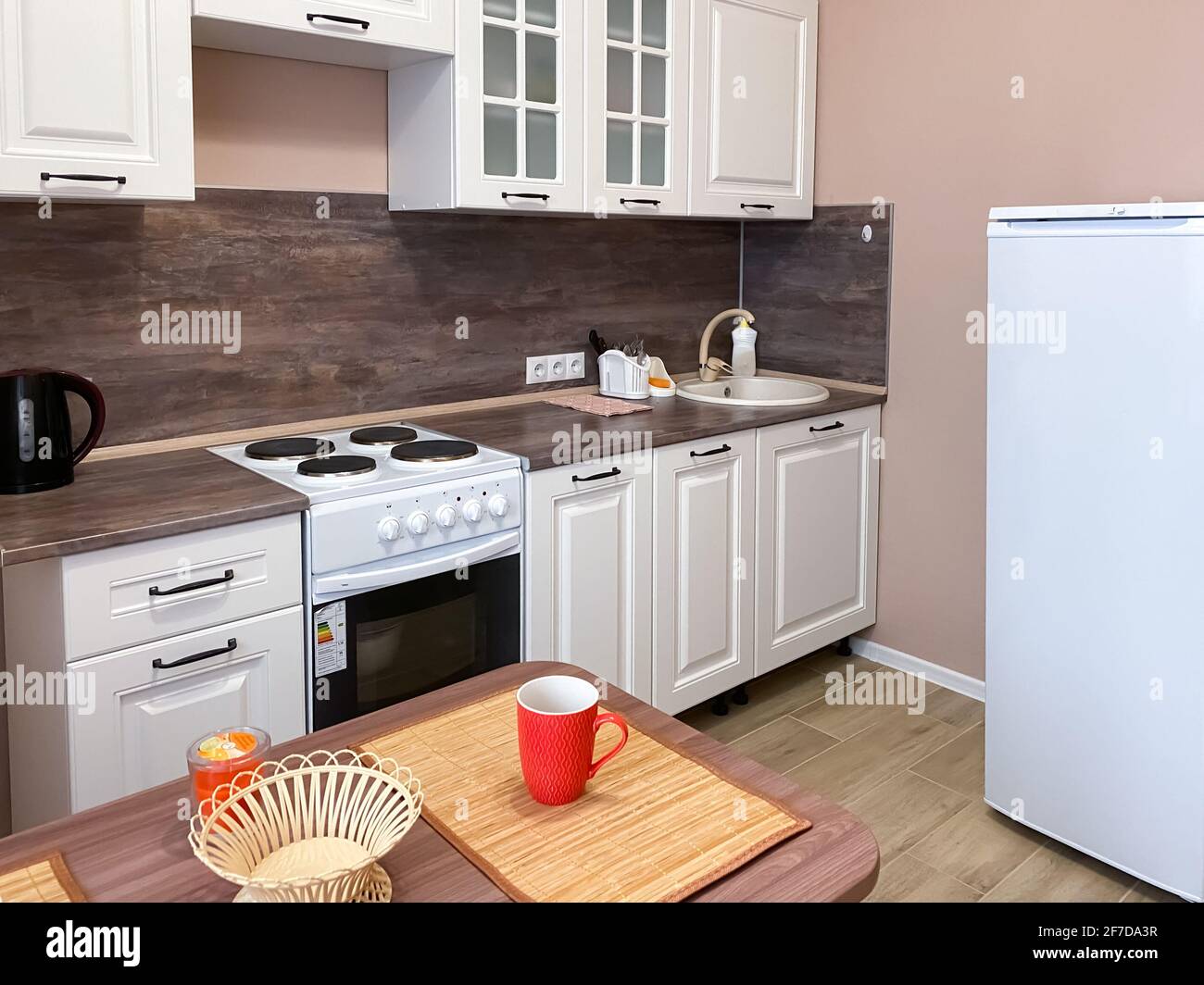 Interior de cocina beige con juego de cocina blanca y electrodomésticos, mesa con utensilios. Foto de stock