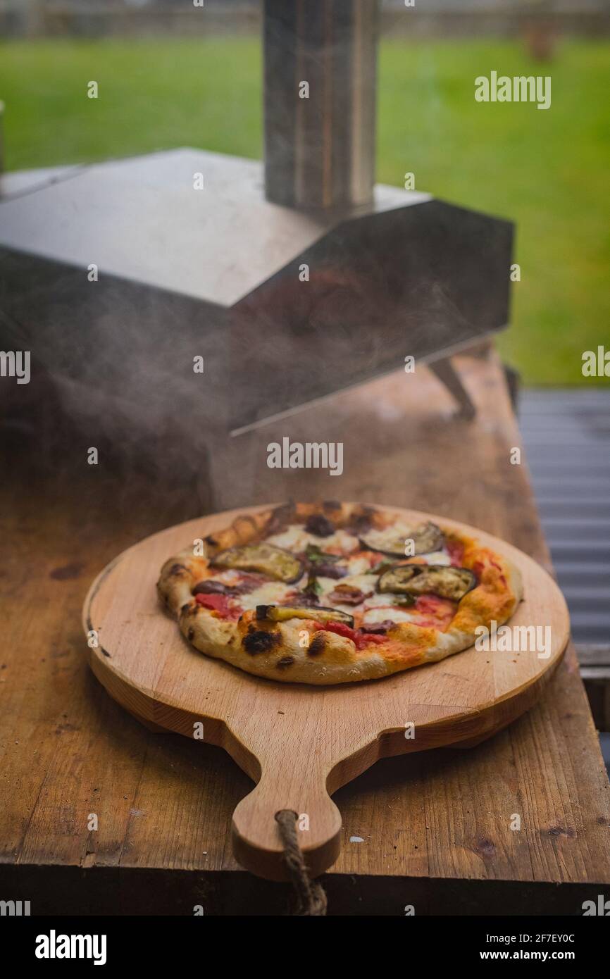 Deliciosa pizza casera fresca en un plato redondo de madera, directamente  de un horno portátil casero de acero inoxidable alimentado por pellets.  Fiesta de pizza al aire libre Fotografía de stock -