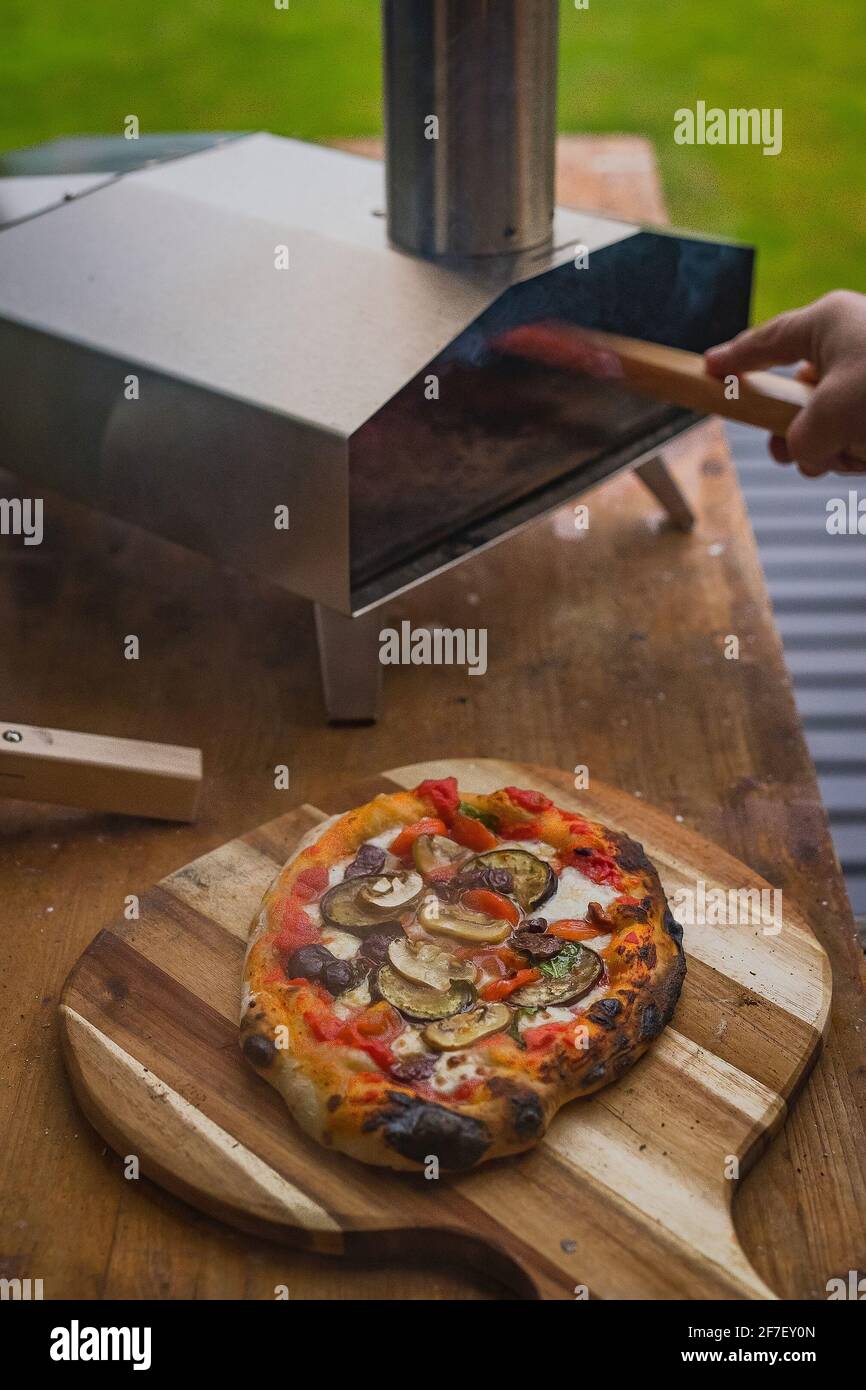 Deliciosa pizza casera fresca en un plato redondo de madera, directamente  de un horno portátil casero de acero inoxidable alimentado por pellets.  Fiesta de pizza al aire libre Fotografía de stock -