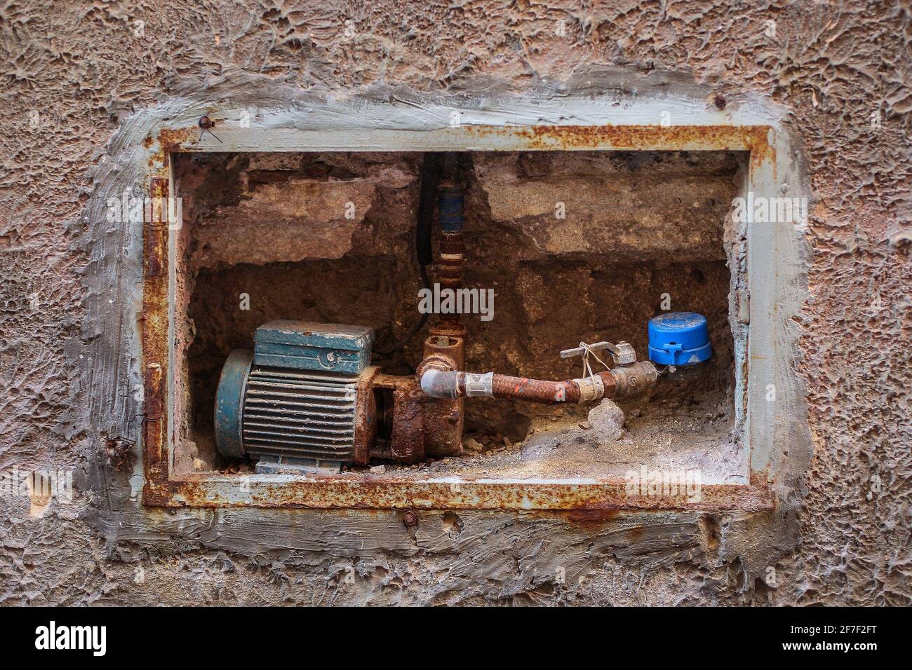 Una bomba de agua de motor eléctrico antigua instalada en una pared de una  casa. Bomba de agua antigua oxidada con tubería visible, válvula y  contador. Concepto de viejo p Fotografía de