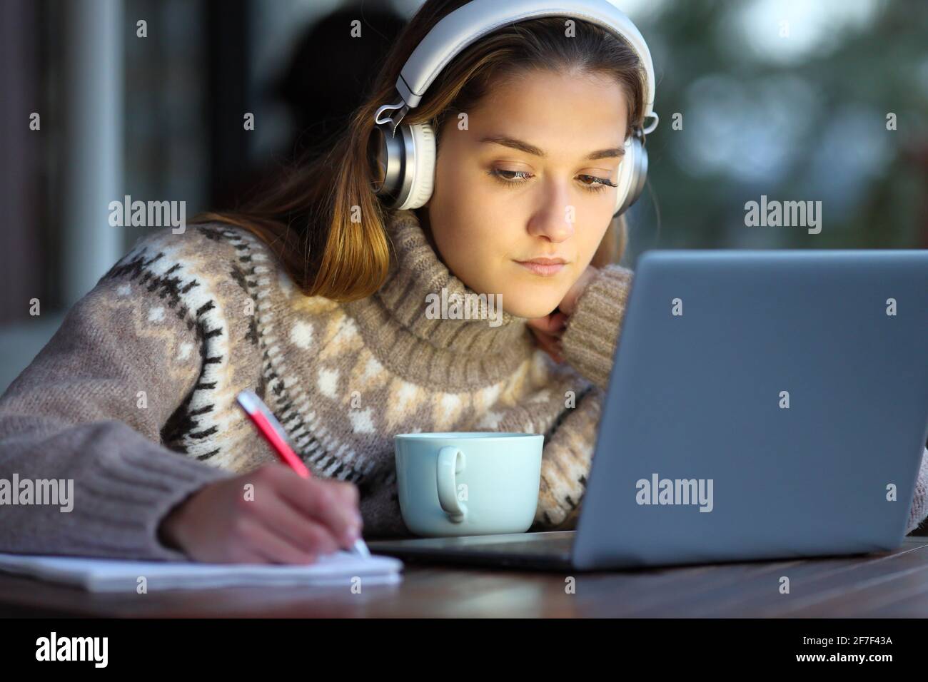 Estudiante concentrado usando auriculares e-learning con portátil tomando notas cuaderno en una cafetería en invierno Foto de stock