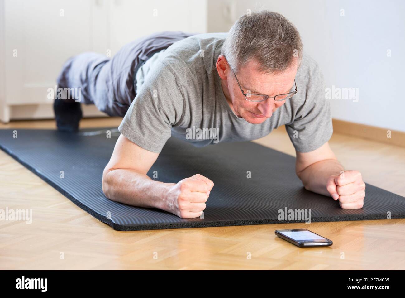 Hombre maduro haciendo ejercicio de la aptitud en el hogar mientras que mira un smartphone: enfoque selectivo en la cara Foto de stock