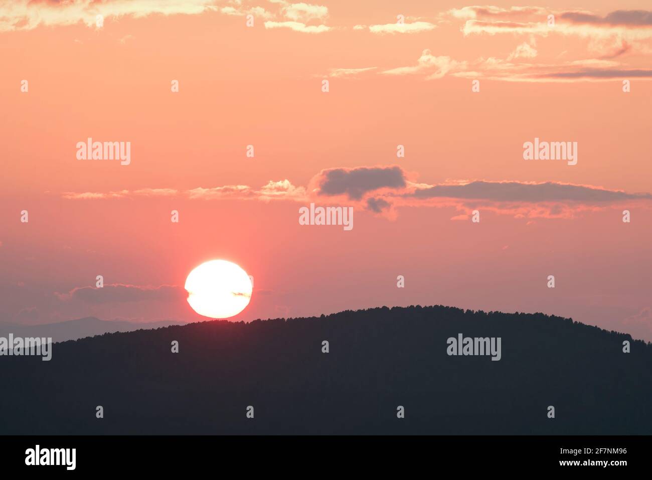 Increíble y delicada puesta de sol rosa con un disco redondo para el sol los contornos de las montañas cubiertas de bosque y nubes iluminadas en el cielo Foto de stock