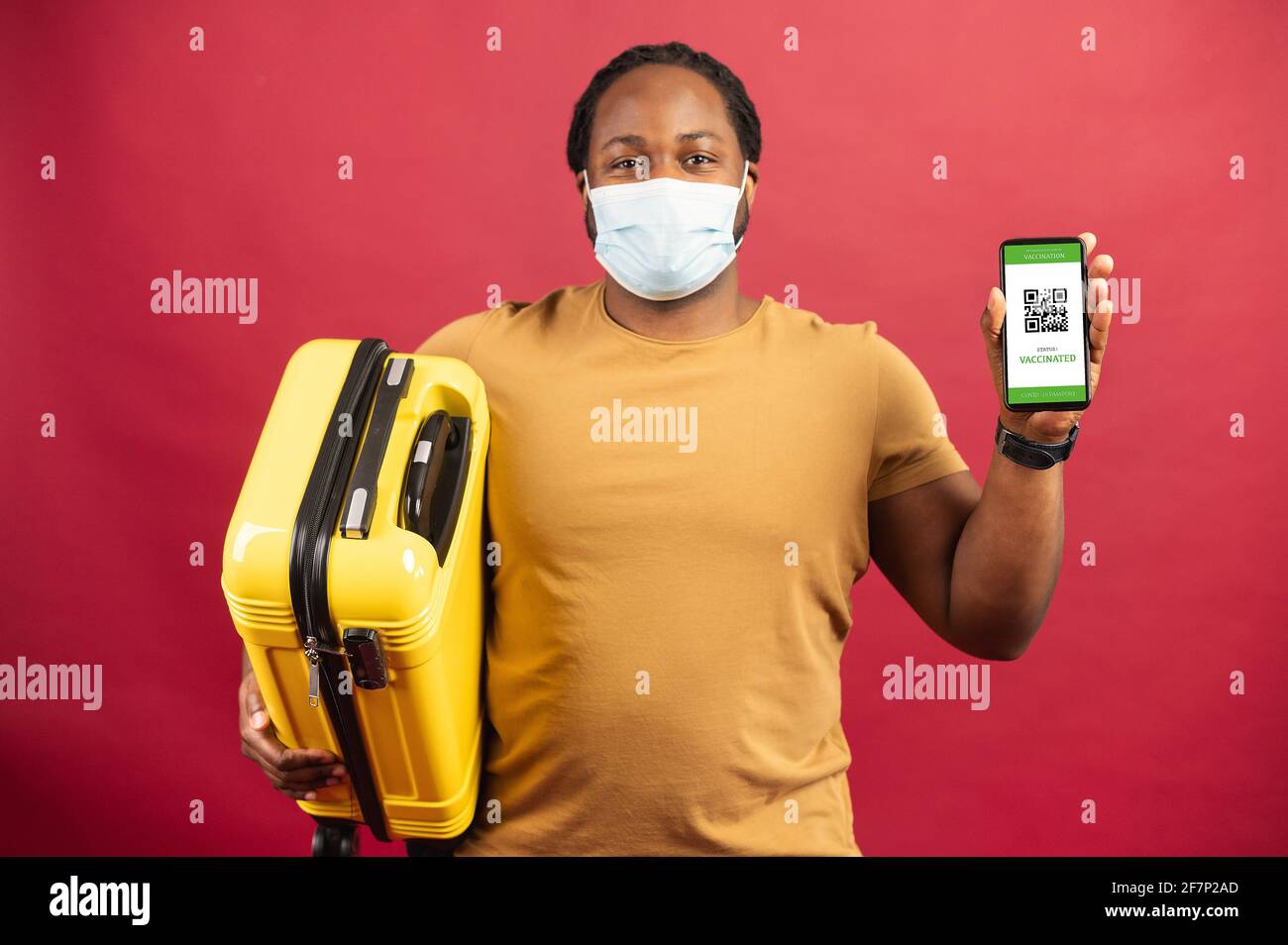 Feliz hombre africano en máscara médica de pie sobre el fondo rojo sosteniendo un bolso amarillo para viajar y celular con código QR en la pantalla, concepto de visualización de boletos sin contacto, paso seguro de aduanas Foto de stock