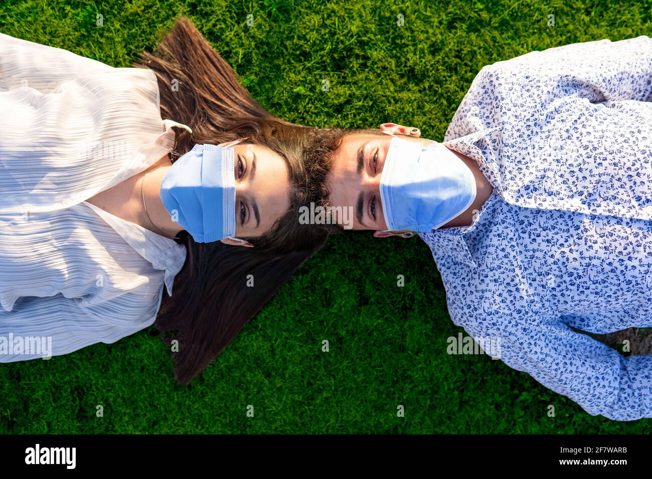 Hermosa pareja de jóvenes estudiantes acostados de cabeza a cabeza está detrás del césped verde del parque de la ciudad con vistas al cámara con la máscara protectora contra Foto de stock