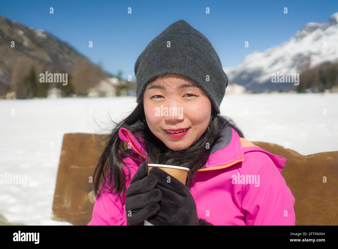 Helando Vacaciones de invierno - jóvenes felices y hermosos chinos asiáticos mujer en el banco en el lago congelado y montañas nevadas bebiendo café o chocolate caliente e Foto de stock
