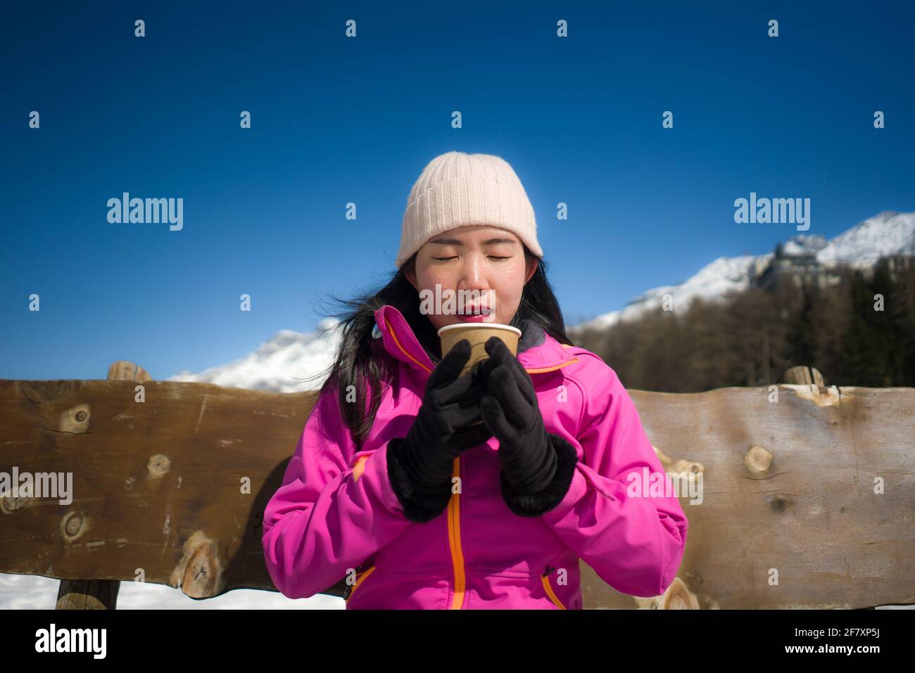 Congelación Vacaciones de invierno - jóvenes felices y hermoso asiático coreano mujer en el banco en el lago congelado y montañas nevadas bebiendo café o chocolate caliente en Foto de stock