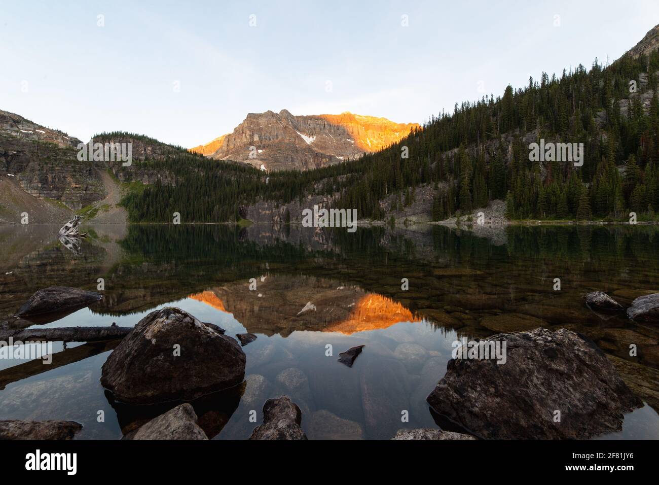 pico de montaña con luz naranja de un amanecer que se refleja en el lago en primer plano con rocas Foto de stock