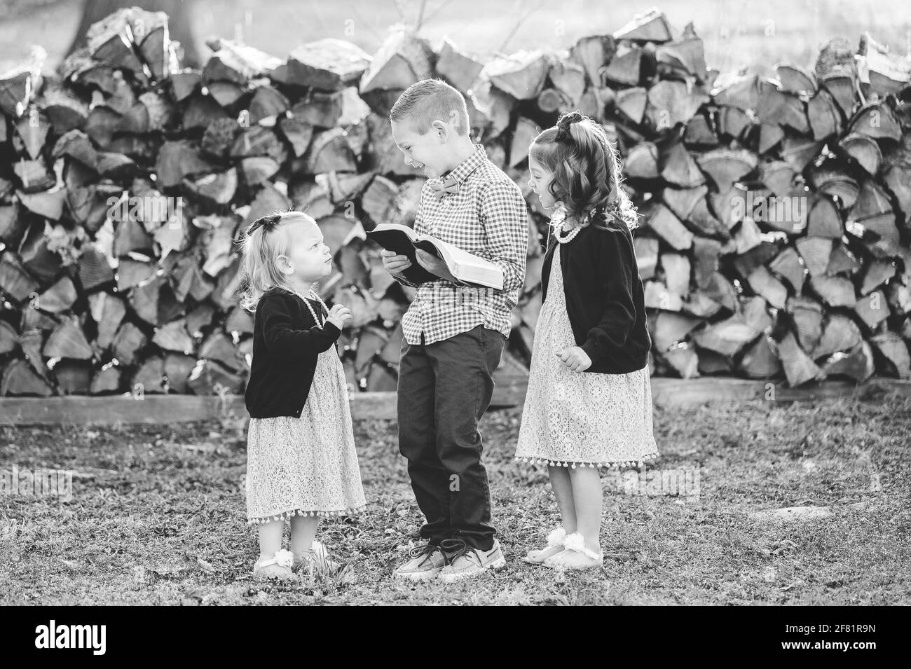 Una fotografía en escala de grises de un niño pequeño leyendo un libro para dos niñas de pie delante de leña apilada Foto de stock