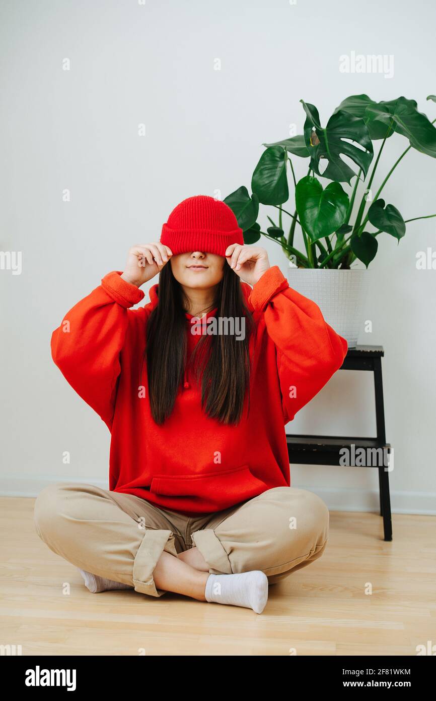 Mujer divertida con ropa de color rojo vivo con la gorra baja sobre sus  ojos. Ella está sentada con sus piernas cruzadas. Ella tiene el pelo largo  recto en lados. Interior Fotografía