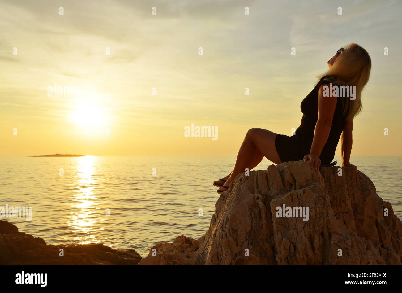 Una mujer sentada en un acantilado está viendo la puesta de sol sobre el mar. Foto de stock