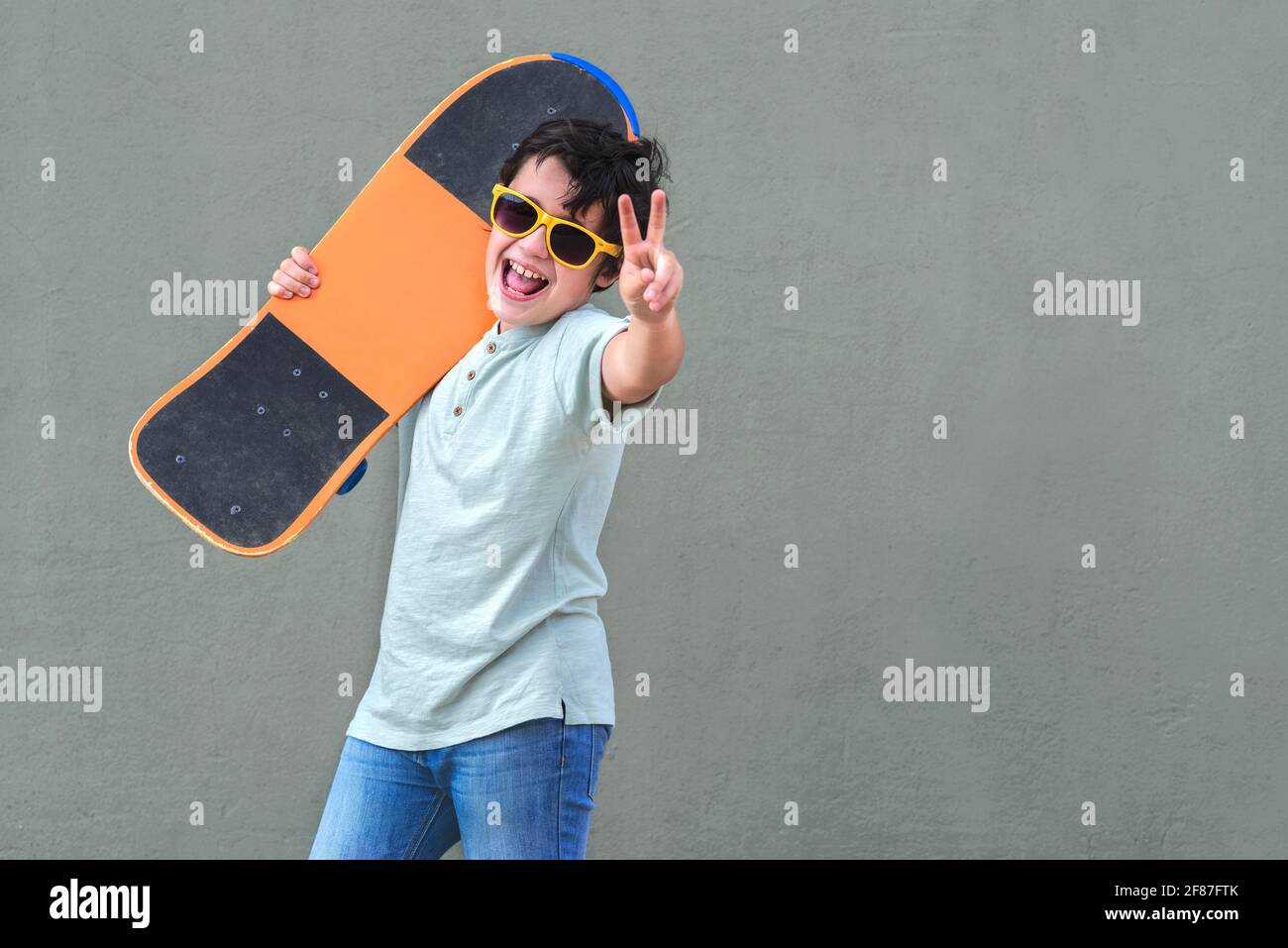 niño feliz y sonriente con skateboard y gafas de sol en el calle sobre fondo verde Foto de stock
