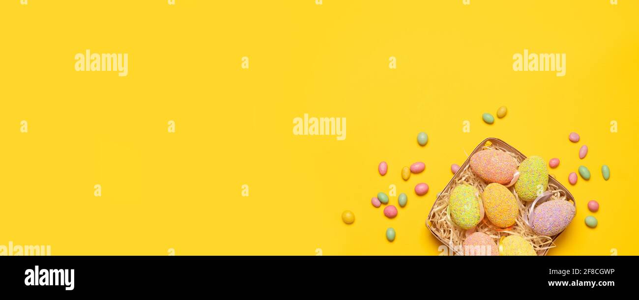 Cesta festiva con huevos de Pascua de colores y sabrosas habas de gelatina de caramelo de colores sobre un fondo amarillo con espacio para copias, vista superior, puesta plana. Horizontal Foto de stock