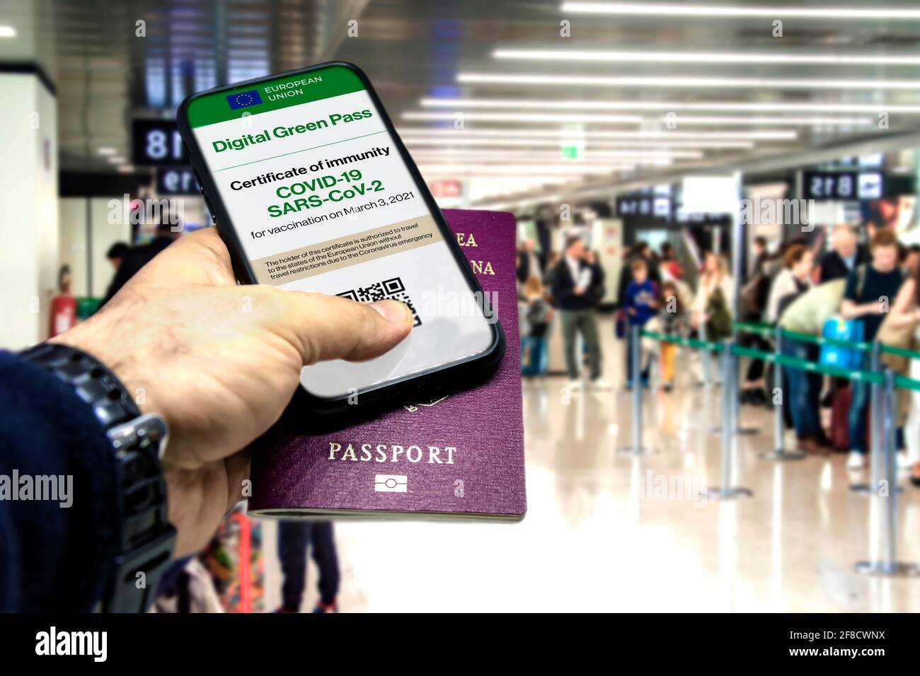 Mano masculina sosteniendo un teléfono con el certificado de vacunación de la Unión Europea en la pantalla y un pasaporte tradicional dentro de un aeropuerto. Inmunidad de Co Foto de stock