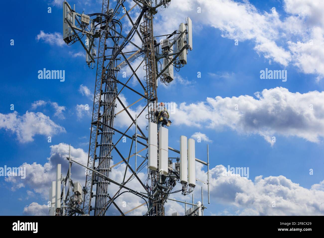 Mantenimiento de telecomunicaciones. Hombre escalador en la torre sobre fondo azul cielo nublado. Estación de mástil de telecomunicaciones, tecnología inalámbrica de antena de TV Foto de stock