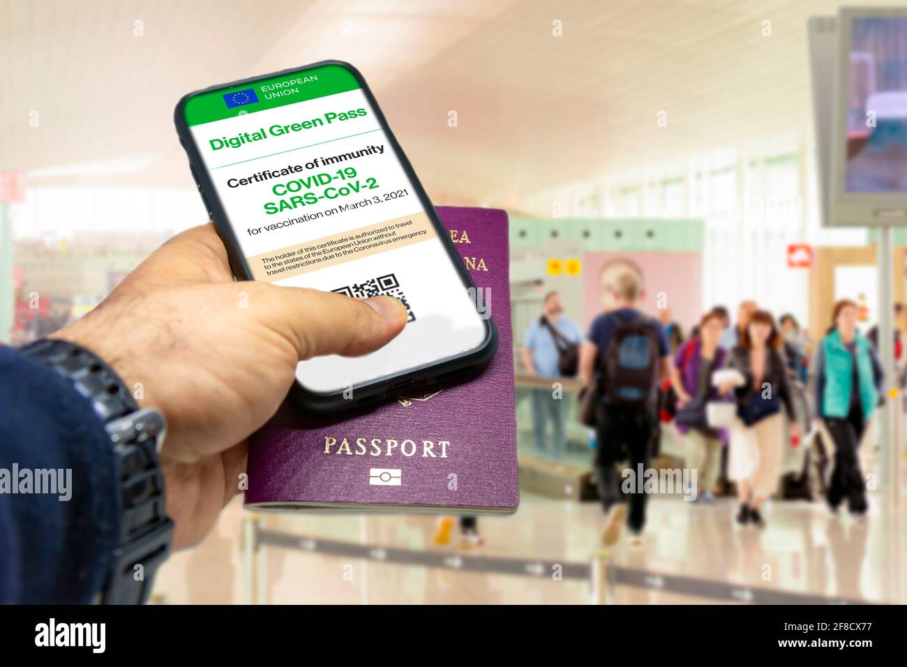 Mano masculina sosteniendo un teléfono con el certificado de vacunación de la Unión Europea en la pantalla y un pasaporte tradicional dentro de un aeropuerto. Inmunidad de Co Foto de stock