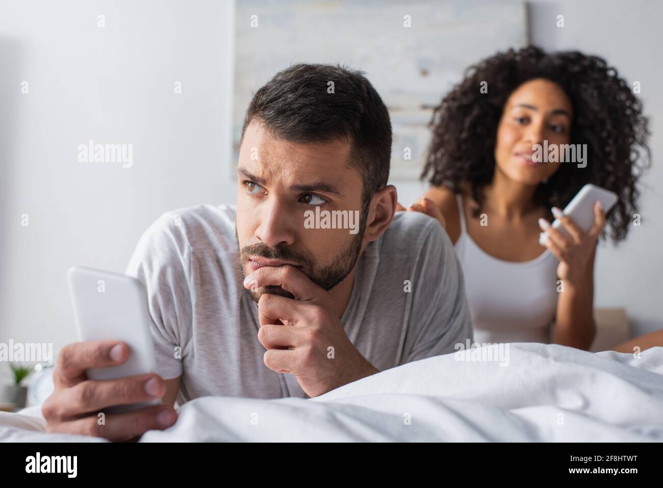 hombre pensativo tumbado en la cama con un smartphone cerca de afroamericanos mujer sobre fondo borroso Foto de stock