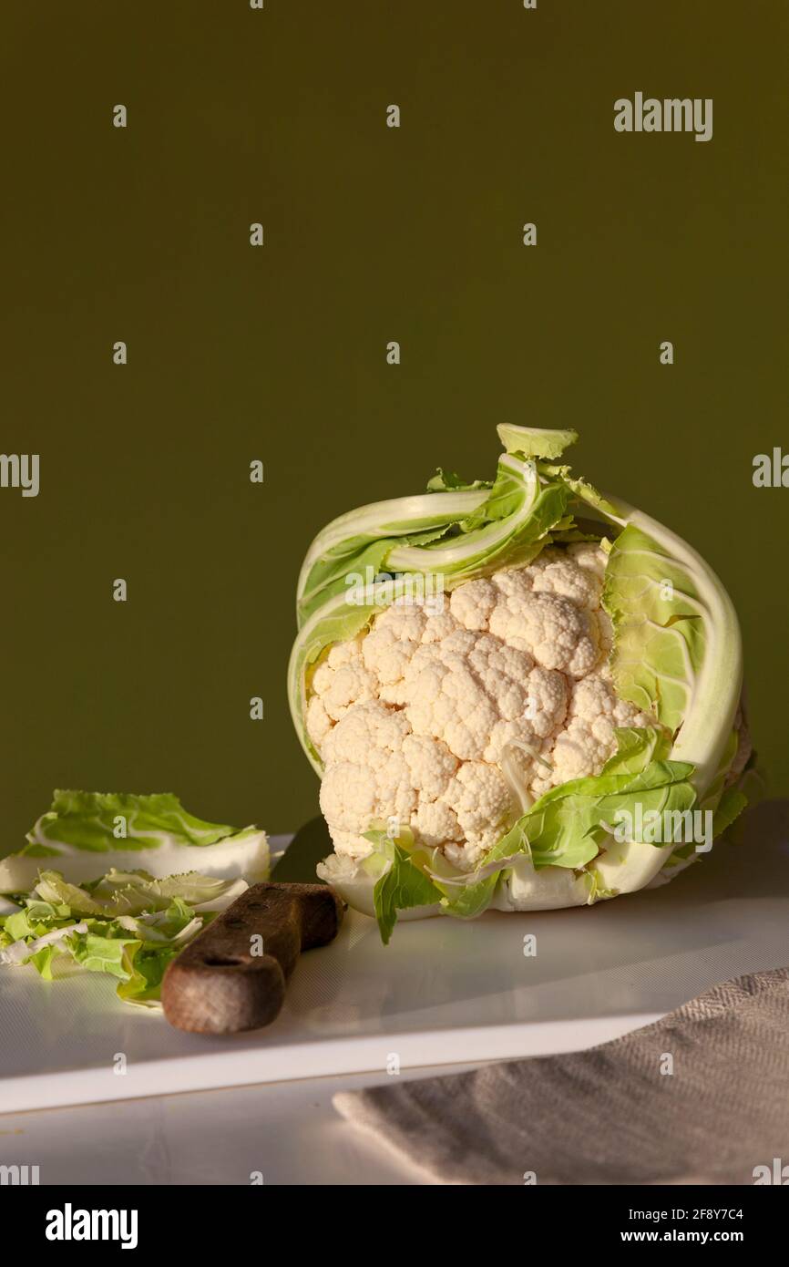 Una coliflor entera sobre una tabla de cortar blanca con cuchillo, servilleta y un fondo verde de color liso Foto de stock