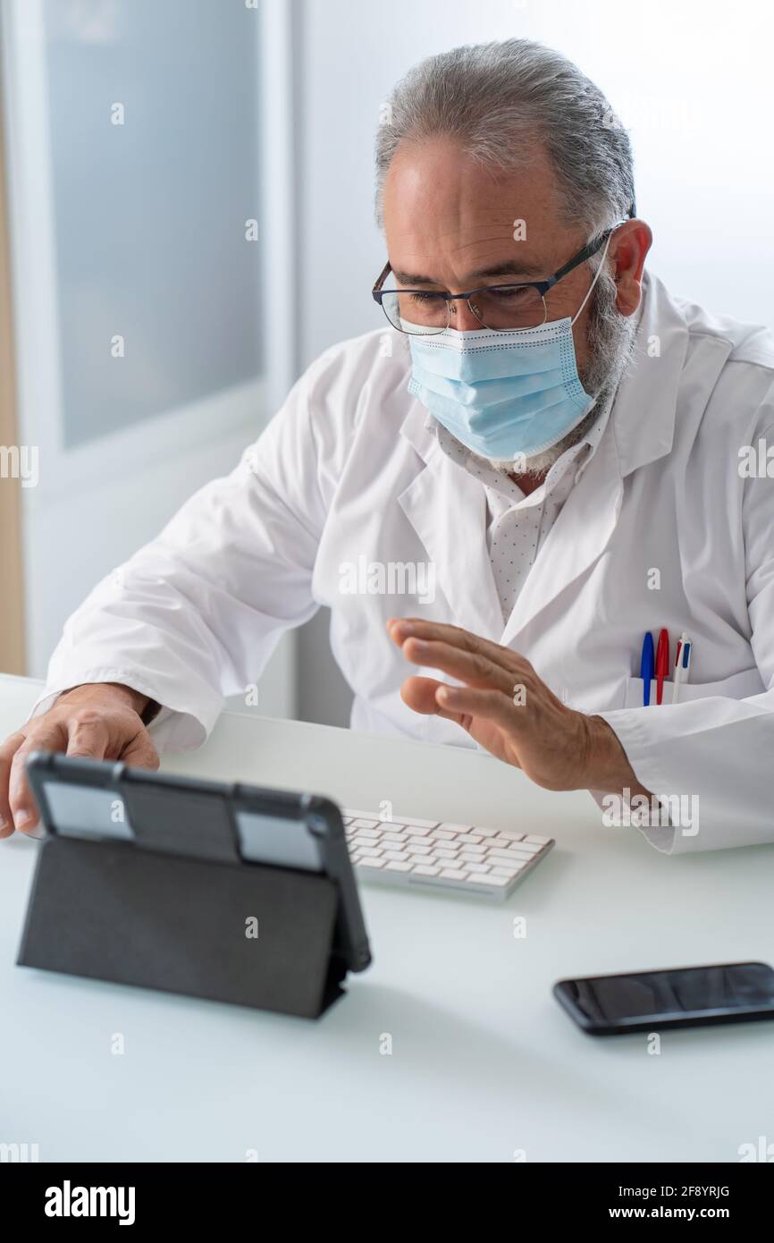 Doctor en abrigo blanco con máscara de saludo en video conferencia en una tableta en su oficina Foto de stock