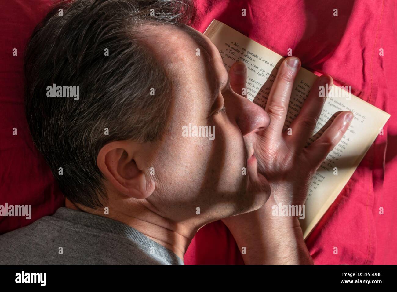 El hombre de mediana edad se durmió con la cabeza en rojo almohada y su mano en un libro abierto con juego de luz y sombra en su rostro Foto de stock