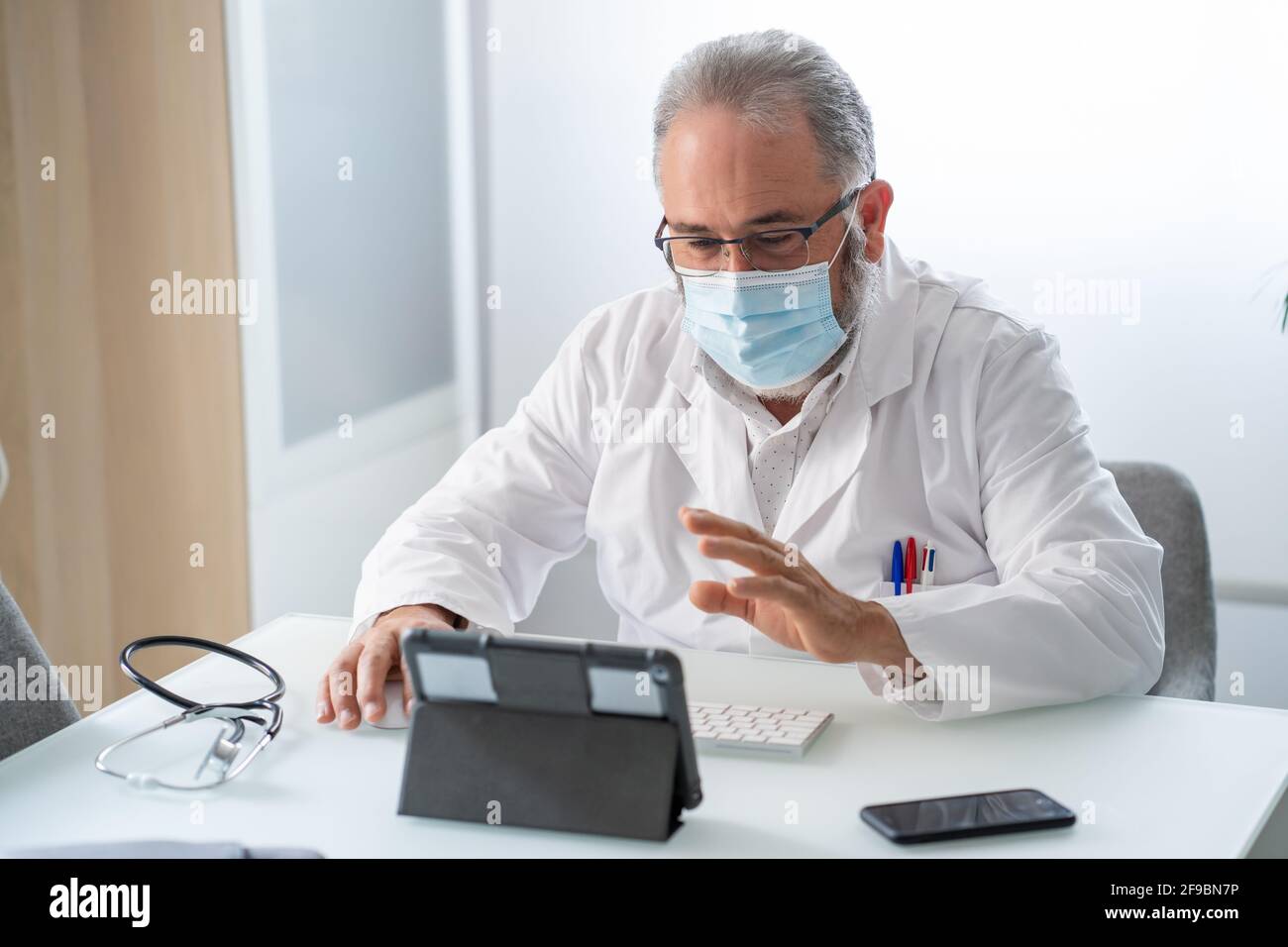 Doctor en abrigo blanco con máscara de cara que tiene un vídeo conferencia con tableta en su oficina Foto de stock