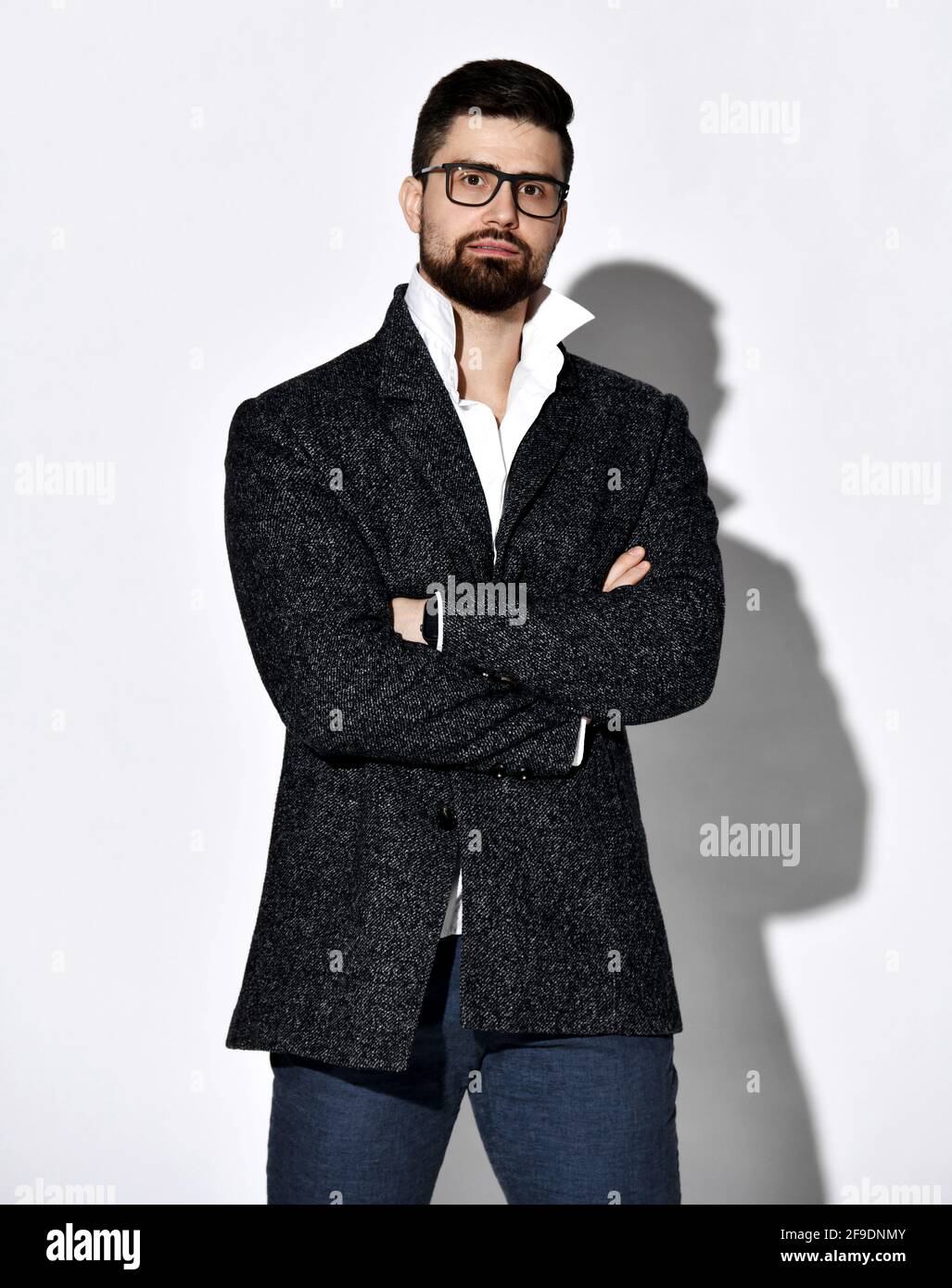 Hombre joven con barba y estilo seguro de sí mismo con gafas y tweed la chaqueta está de pie con las manos cruzadas en el pecho Foto de stock