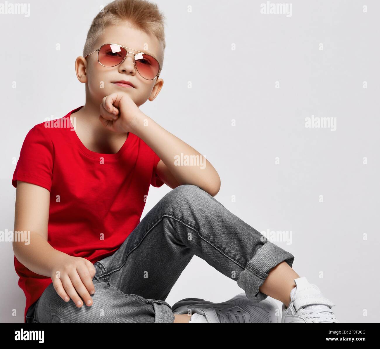 Alegre y elegante chico con camiseta roja, jeans, sneakers y gafas de sol  sentado en el suelo sosteniendo la barbilla con la mano mirando la cámara  Fotografía de stock - Alamy