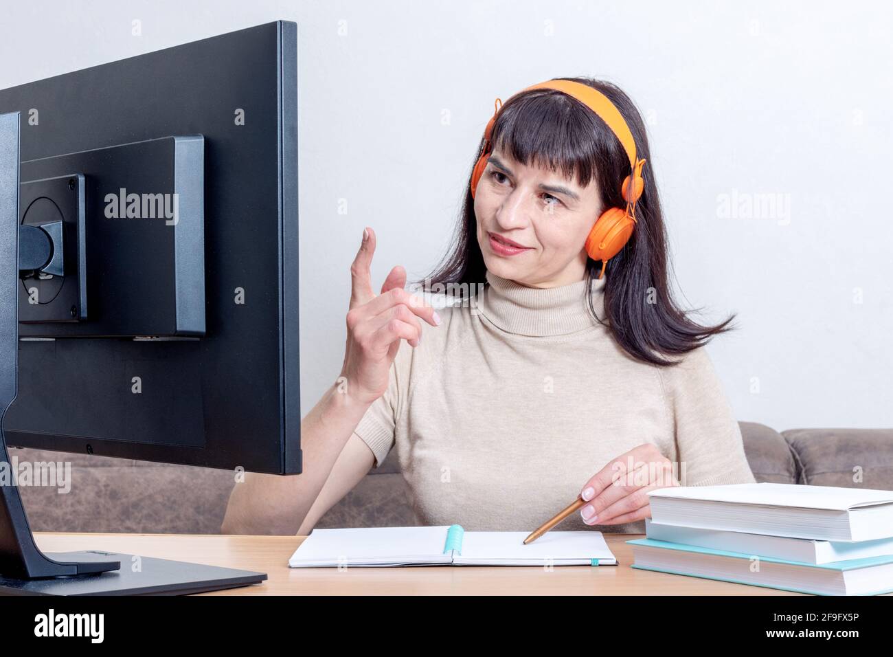 Atractiva mujer con auriculares de color naranja sentada en una mesa con  libros, mirando un monitor de ordenador y levantando el dedo, charlando en  Internet Fotografía de stock - Alamy