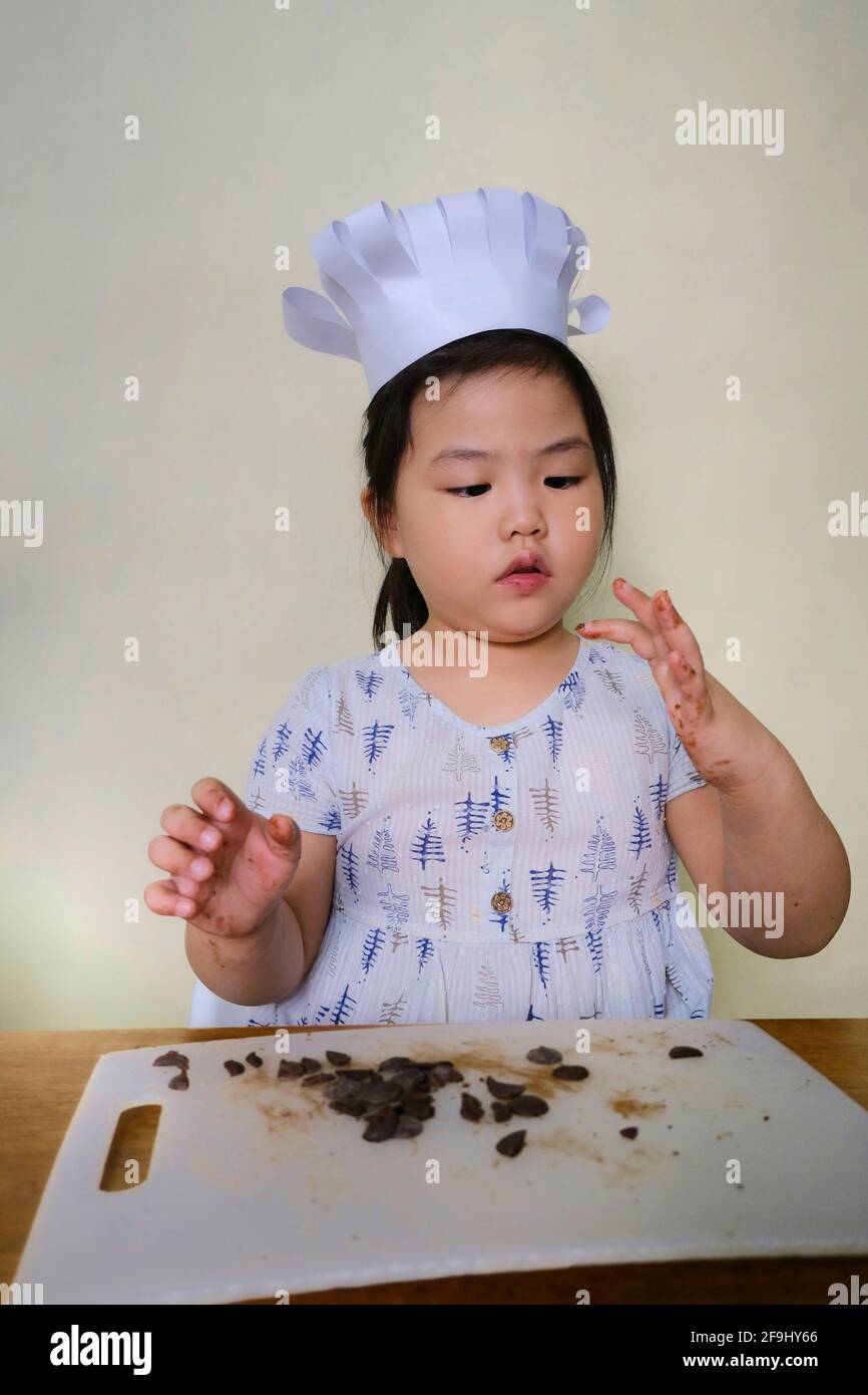 Una chica asiática joven y linda con un sombrero de chef blanco está aprendiendo a cocinar con su madre, picando chocolate y degustándolo antes de mezclarlo en el Foto de stock