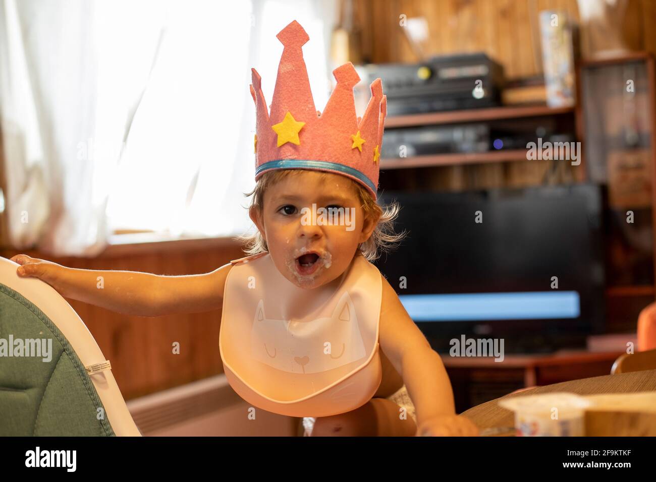 Adorable niño en una corona se sienta en casa en un babero. Pequeña princesa con gracioso expresión interior de casa Foto de stock