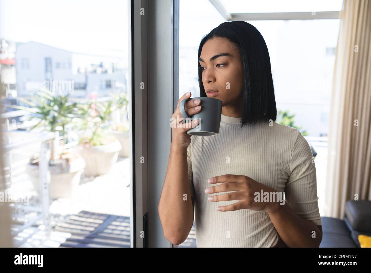 Mujer transgénero de raza mixta de pie mirando por la ventana de la sujeción taza de café Foto de stock