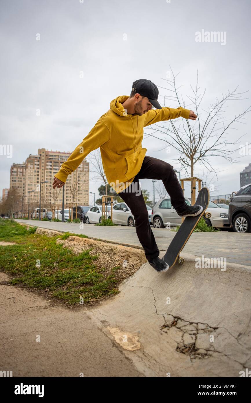 Macho caucásico joven con capucha amarilla y gorra negra saltando con su skate Foto de stock