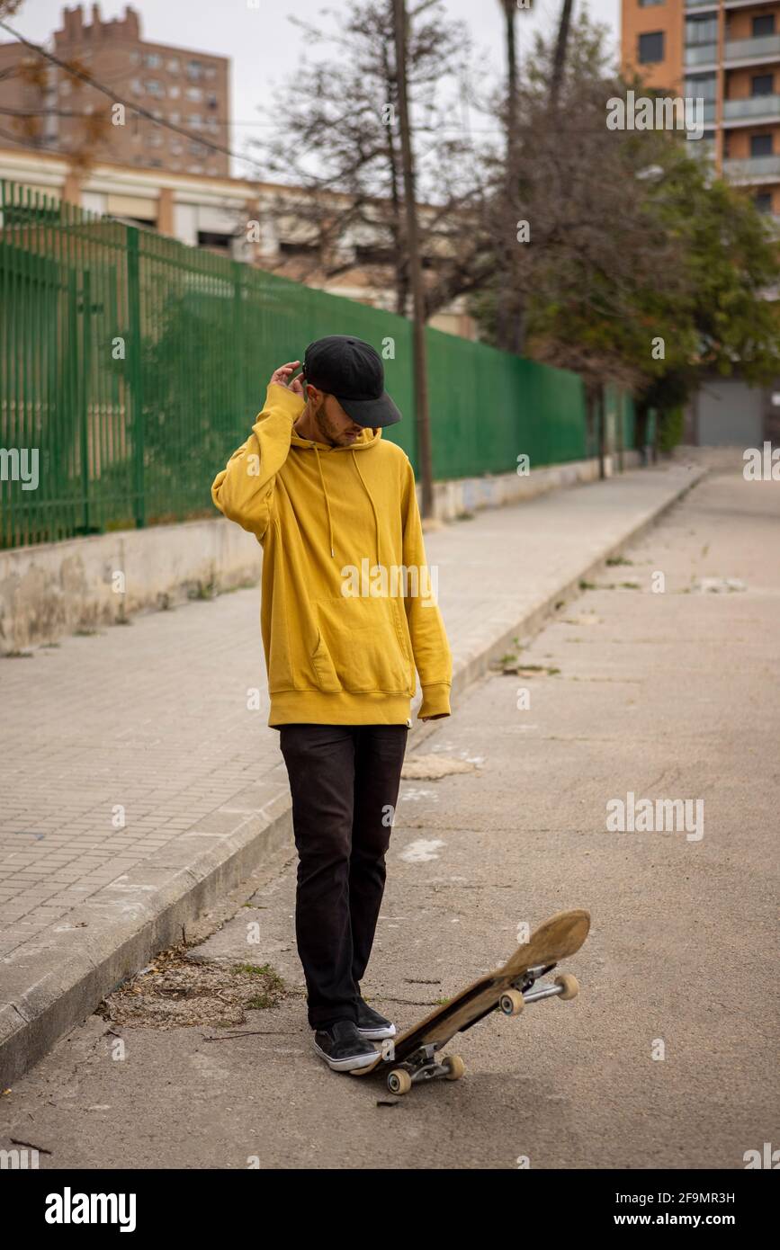 Macho caucásico joven con capucha amarilla y gorra negra de pie sobre su monopatín Foto de stock
