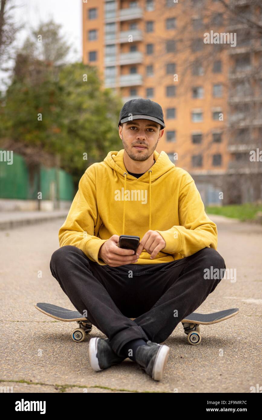 Macho caucásico joven de Italia en una sudadera con capucha amarilla y. gorra negra sentada sobre una tabla de skateboard con un smartphone Foto de stock
