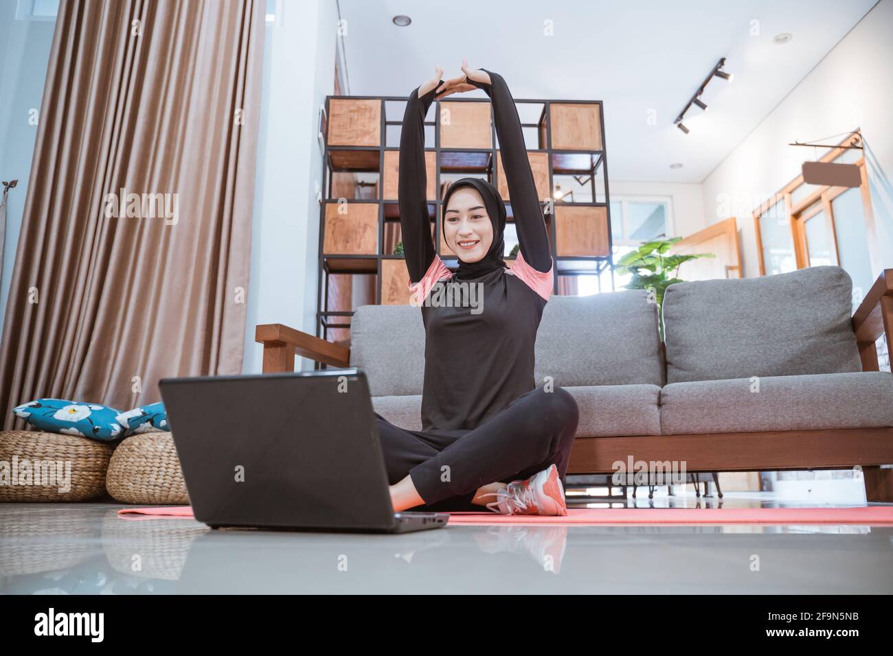 una mujer en un hijab sportswear se sienta con las piernas cruzadas mientras se estira con sus manos hacia arriba mientras mira un portátil Foto de stock