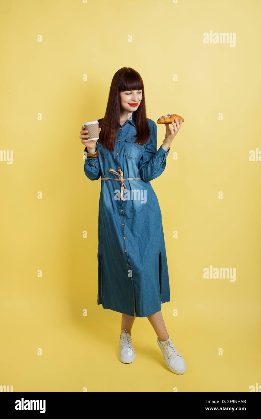 Anuncio promocional de comida y bebida. Tiro completo de dama joven en  informal moderno vestido jeans y sneakers, posando sobre fondo amarillo y  alberga deliciosos croissant y café para llevar Fotografía de