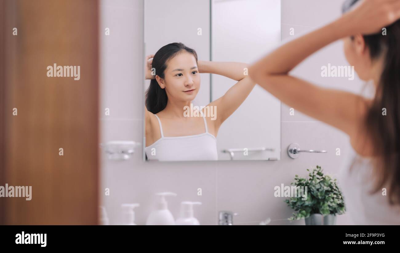 Joven pelirroja mujer atando su cabello en un moño delante del espejo en un baño vista desde la parte trasera Foto de stock