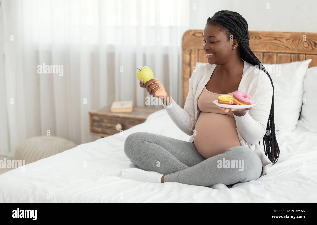 Nutrición prenatal. Mujer negra elegir entre manzana y donuts en casa de stock Alamy
