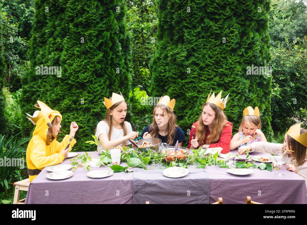 Los niños lloraban coronas de papel sentados en una mesa decorada comiendo a la parrilla salchichas celebrando la fiesta de cumpleaños en un jardín verde Foto de stock