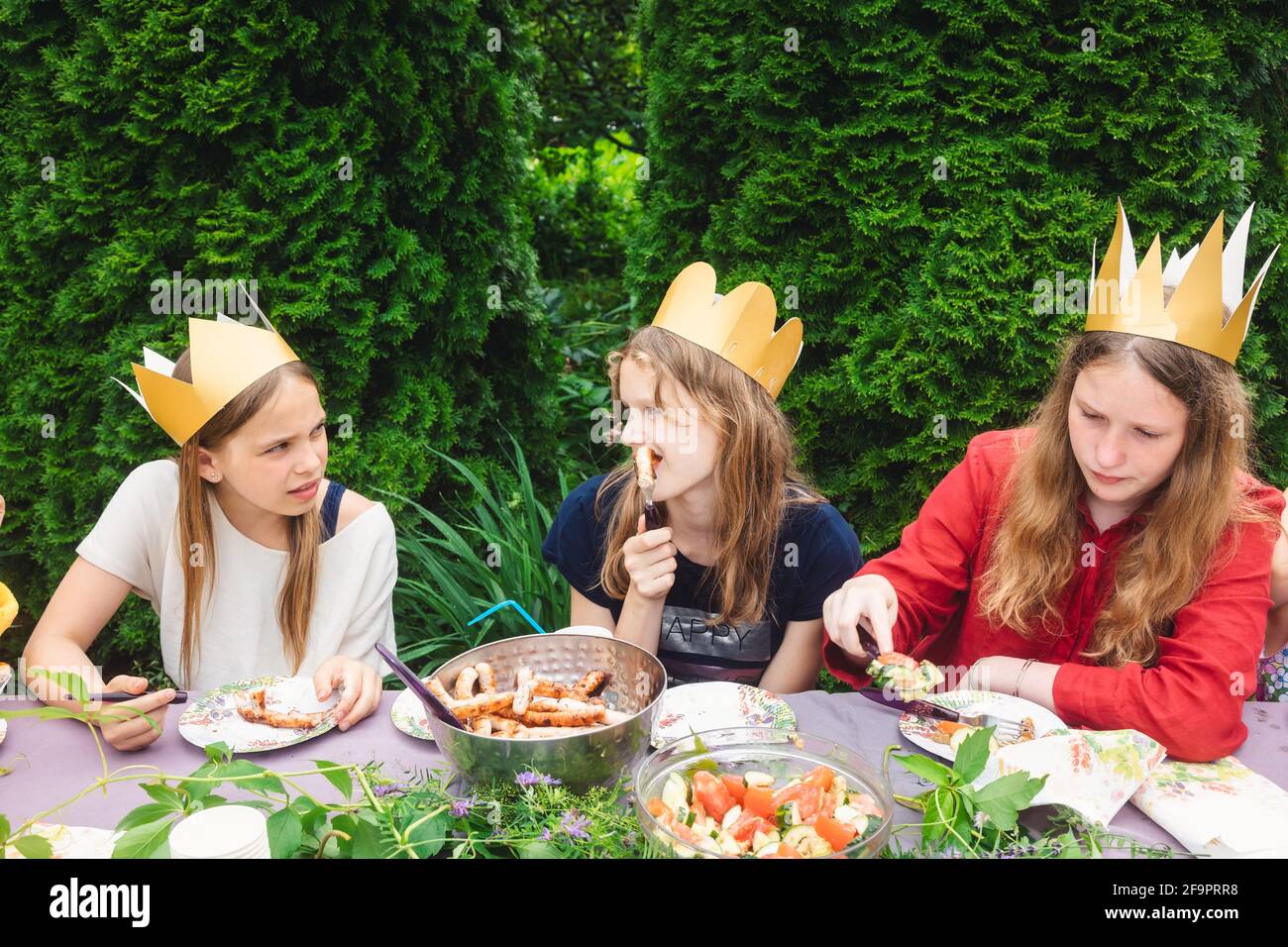 Los niños lloraban coronas de papel sentados en una mesa decorada comiendo a la parrilla salchichas celebrando la fiesta de cumpleaños en un jardín verde Foto de stock