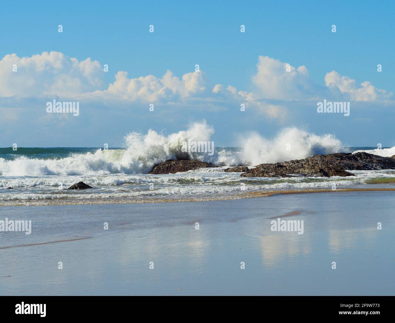 Día de cielo azul en la playa con olas rompiendo sobre las rocas cerca de la orilla frente a nubes blancas reflejadas en la arena mojada, Sawtell Australia Foto de stock