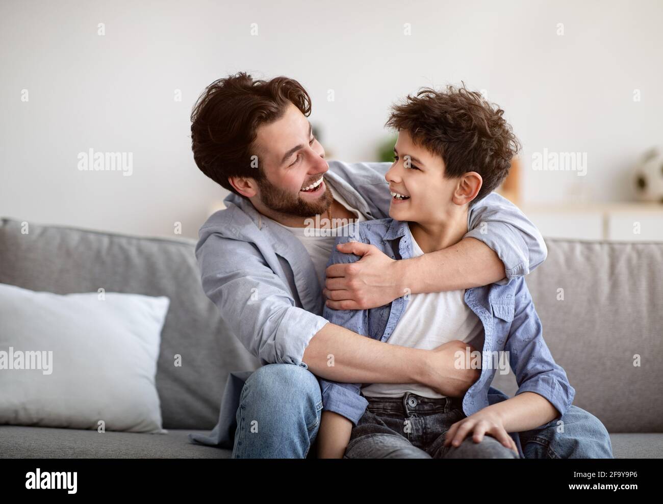 Concepto de dos generaciones. Feliz padre abrazando a su hijo, mirándose el uno al otro y sonriendo, sentado en el sofá en casa Foto de stock