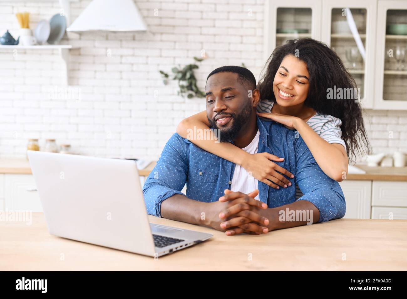 Mujer biracial abrazando al hombre de detrás, pareja étnica mixta relajarse en el nuevo apartamento en casa navegando por Internet en el portátil, mostrar el amor todos los días de las maneras más simples, viendo vídeo, mirando la pantalla Foto de stock