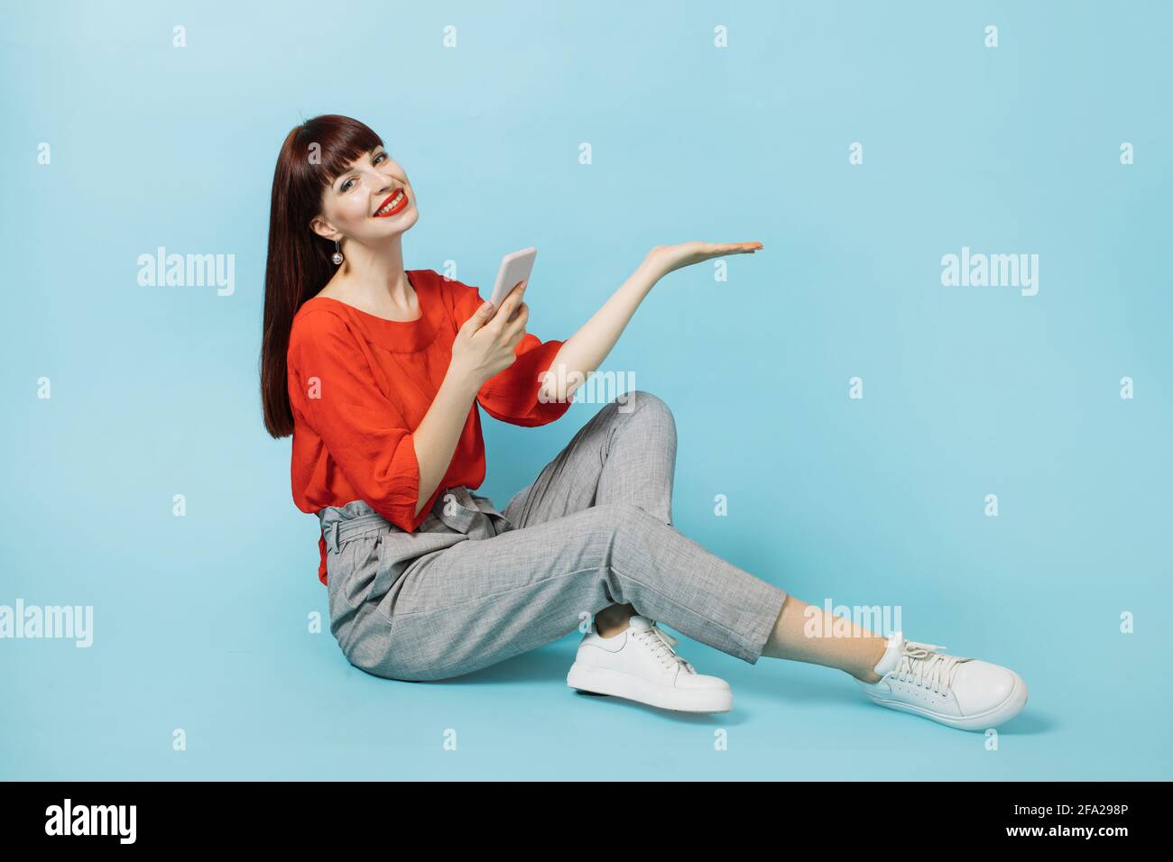Señorita joven en ropa elegante casual, sosteniendo el teléfono, mostrando  algo imaginario, vacío espacio de copia para algún texto, producto o  eslogan, en la palma de la mano abierta, sentado aislado sobre