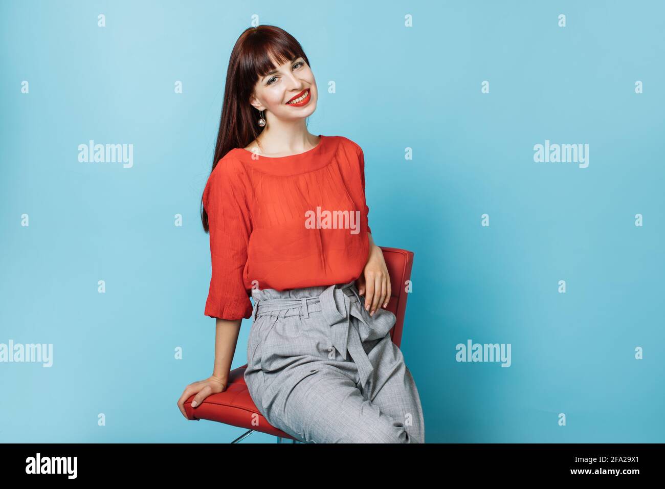 Cintura para arriba retrato de hermosa mujer de pelo rojo de 30 años, con  camisa de moda roja y pantalones grises, sonriendo, sentado en una silla  roja y relajante. Mujer posando sobre