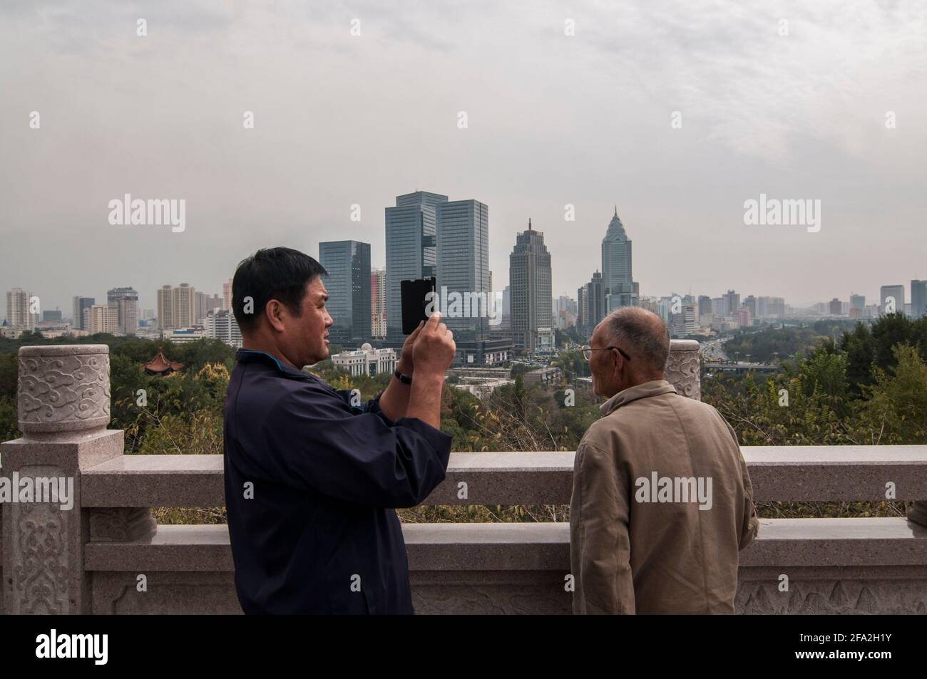 Un hombre toma una foto con su teléfono de otro hombre frente a una vista del horizonte con oficinas, edificios residenciales de gran altura y rascacielos del distrito central de Jiefang en Urumuqi en un día de nebly en Xinjiang, China, PRC. © Capturas de tiempo Foto de stock