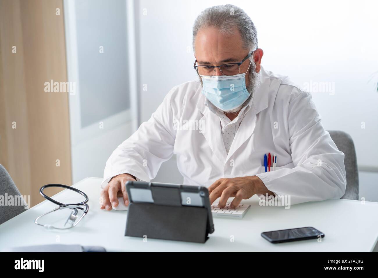 Doctor con mascarilla Tener una videoconferencia con tableta en su oficina Foto de stock