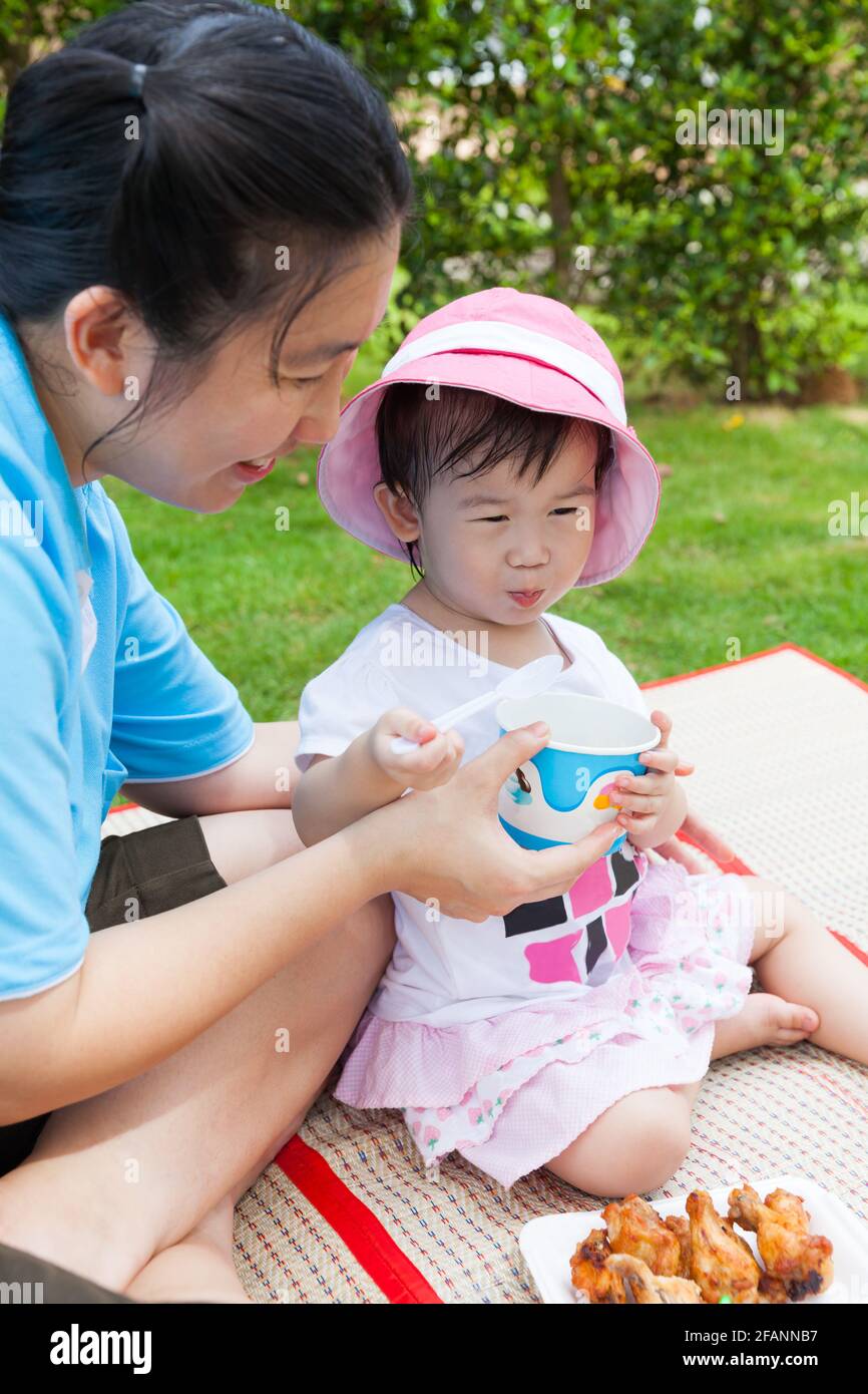 Vacaciones, madre y adorable niña asiática (tailandesa) disfrutar de su almuerzo en el parque, niño sosteniendo cuchara y taza, pollo frito en el plato, más Foto de stock