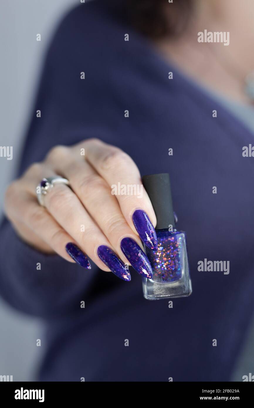 Mano femenina con uñas largas y manicura lila púrpura contiene una botella de esmalte de uñas Foto de stock