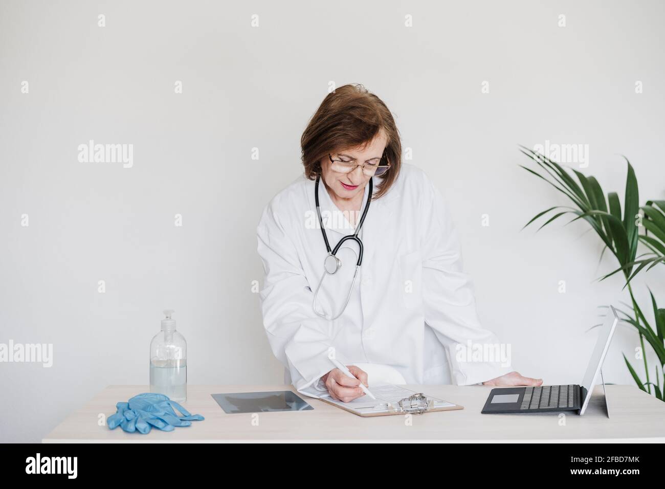 Mujer doctora con tableta digital escribiendo en papel en el portapapeles en el escritorio del hospital Foto de stock